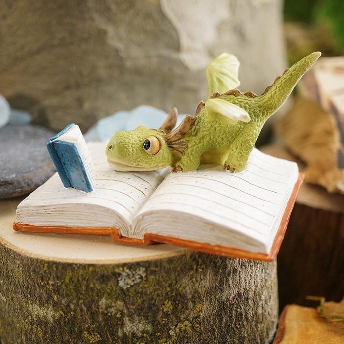 A small green dragon reads a tiny book while lying on an open notebook placed on a wooden surface outdoors.