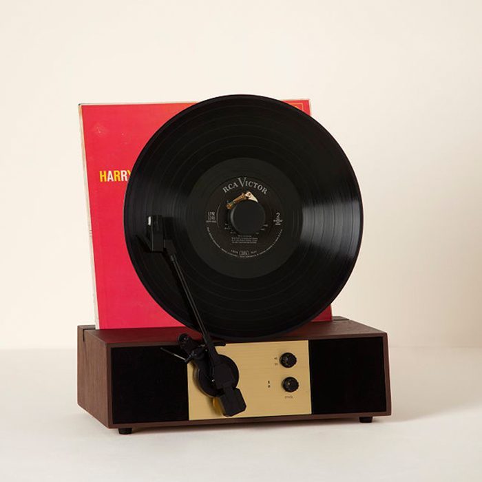 Record player displays upright vinyl rotating; set against a beige background with a red album cover partially visible behind it.
