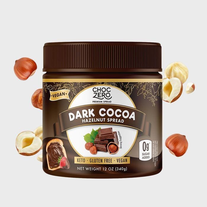 A jar of dark cocoa hazelnut spread rests against a light background, surrounded by hazelnuts. Text highlights its vegan, keto, gluten-free qualities.