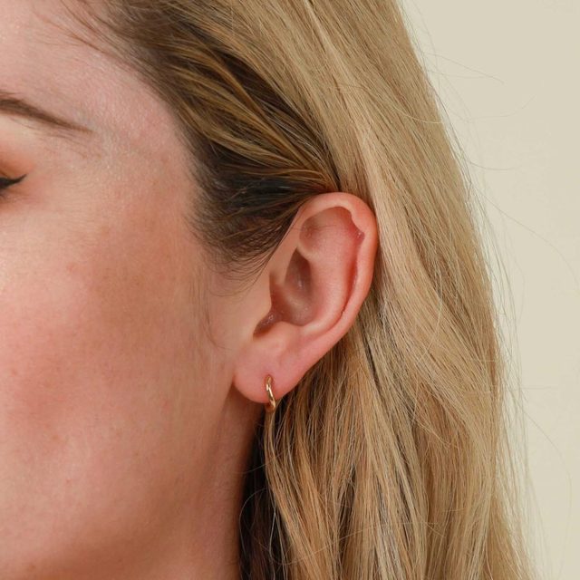 An ear adorned with a small gold hoop earring, surrounded by light brown hair and a neutral background.