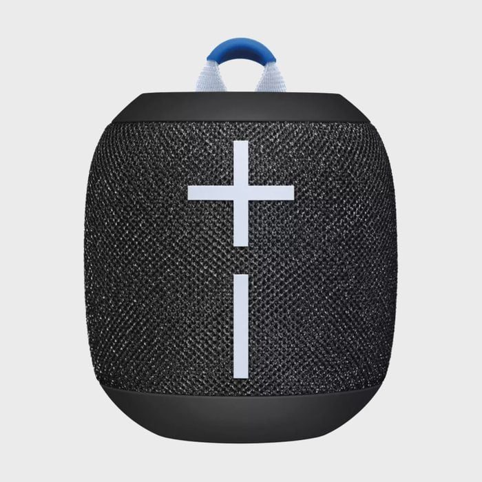 A cylindrical black speaker with a textured surface displays large, white volume control symbols. It features a blue loop on top for carrying.
