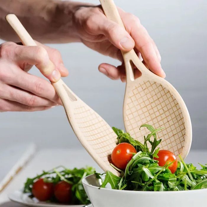 Wooden salad servers lift greens and cherry tomatoes from a white bowl in a kitchen setting.