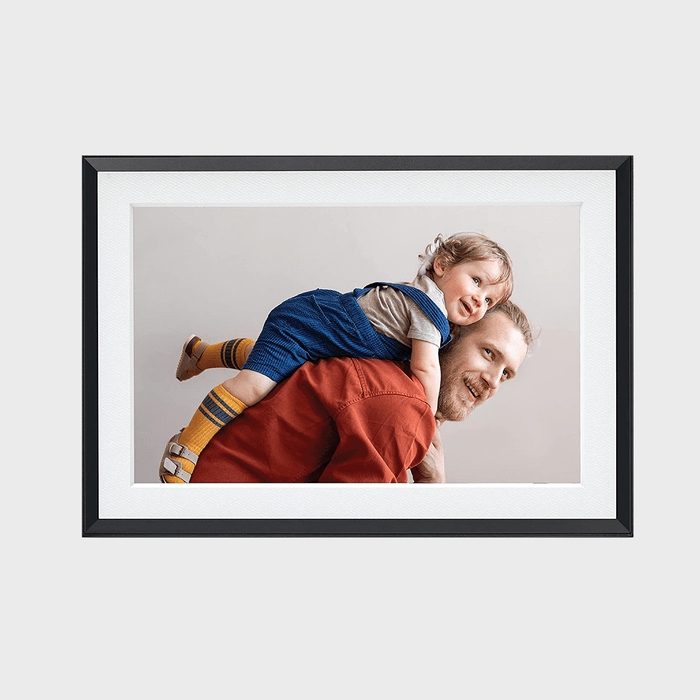 A child rides piggyback on a smiling man, both in colorful clothing, set against a plain background within a black-framed photograph.