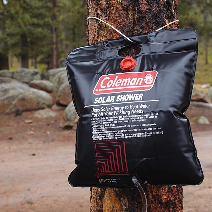 Black solar shower bag hangs on a tree, providing portable heated water in a forested outdoor setting. Text: 