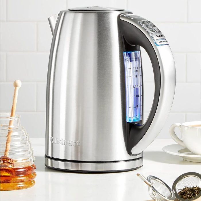 Stainless steel electric kettle sits on a kitchen counter beside a honey jar, strainer, and a teacup, against a tiled wall.