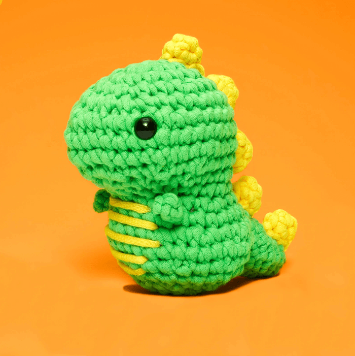 A green crocheted dinosaur toy stands upright with yellow spikes, set against a bright orange background.