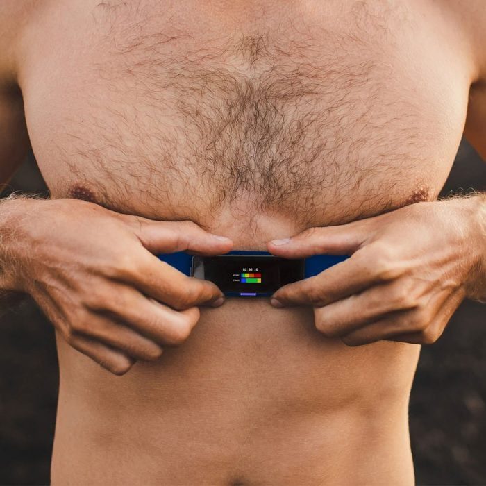 Person holds a small device showing colored bars against a bare chest, outdoors.