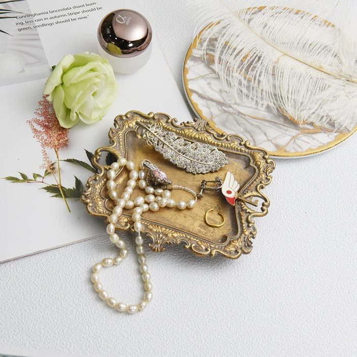 Ornate tray holds jewelry against a textured white surface; surrounded by a decorative plate, flower, magazine, and a small, round container.