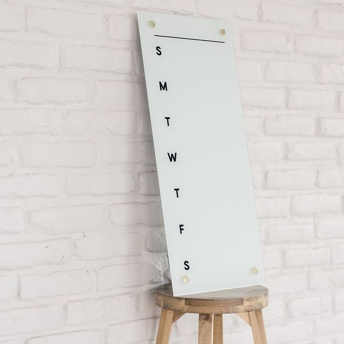 A white board with weekday initials, leaning against a light brick wall, resting on a wooden stool.
