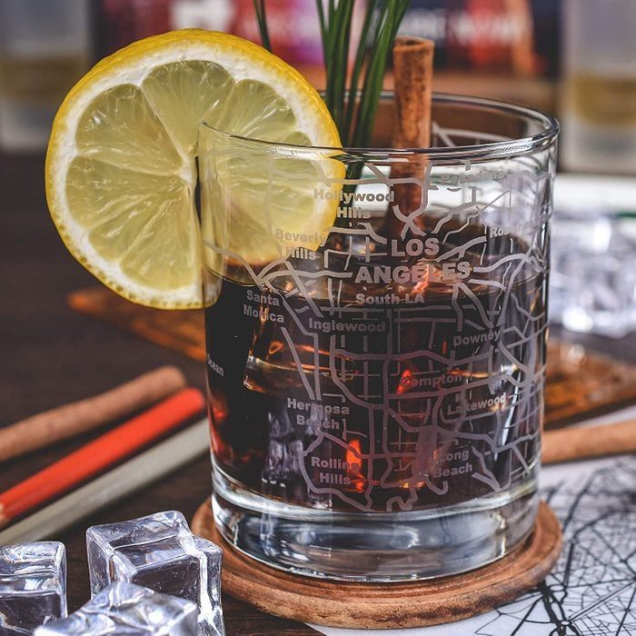 Glass with etched city map holds dark drink, lemon slice, and cinnamon stick. Ice cubes nearby on a table.