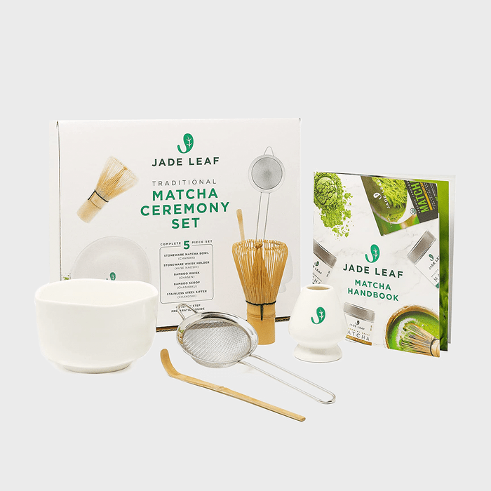 Matcha ceremony set displayed; tools arranged neatly. Includes bowl, whisk, scoop, sifter, holder; packaging and handbook in background. Text: 