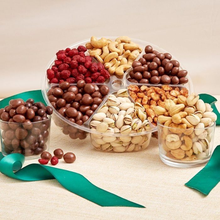 Assorted nuts and chocolates neatly arranged in a circular tray, accompanied by two small clear jars on a beige surface with green ribbon.