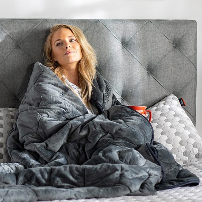 Person relaxes under a gray blanket, holding a red mug, leaning against a tufted headboard in a cozy bedroom.