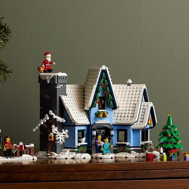 Lego house sits festively decorated, featuring Santa on chimney, surrounded by trees and gifts on a wooden table against a green wall.