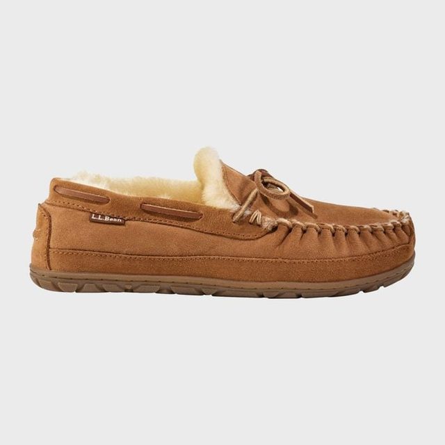 A brown moccasin-style slipper sits with visible fleece lining, in a plain, light gray background. Text reads: 