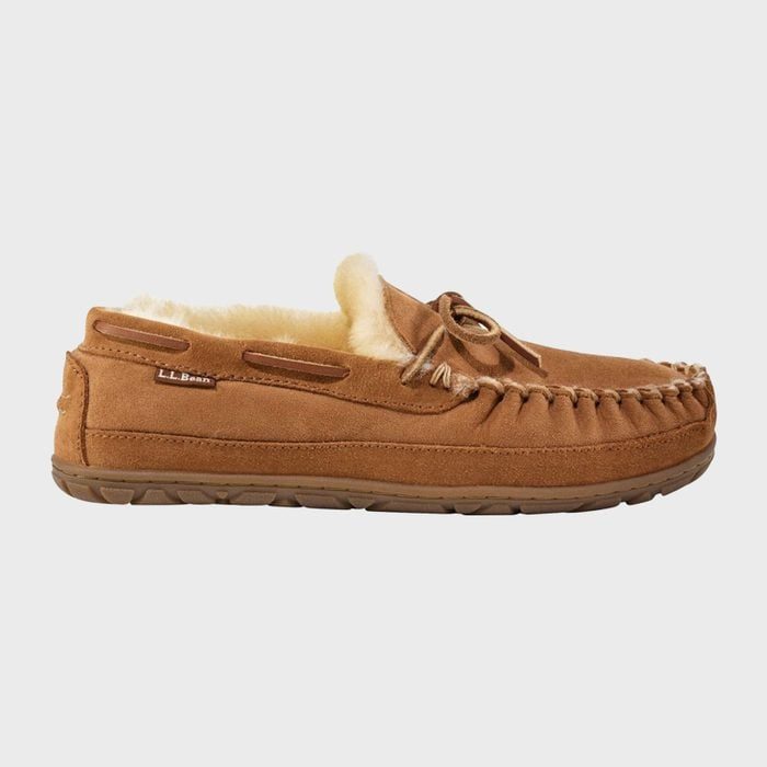 A brown moccasin-style slipper sits with visible fleece lining, in a plain, light gray background. Text reads: 