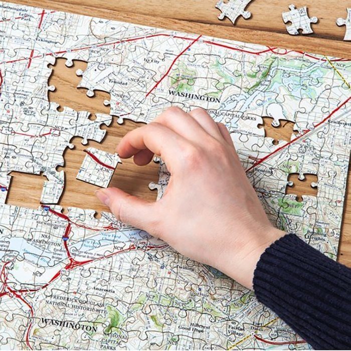 A hand places a map-themed puzzle piece in a partially completed puzzle on a wooden surface.
