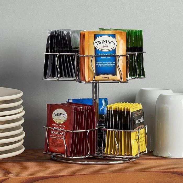 Tea packets rest in a rotating metal holder, surrounded by stacked plates and white mugs on a wooden countertop.