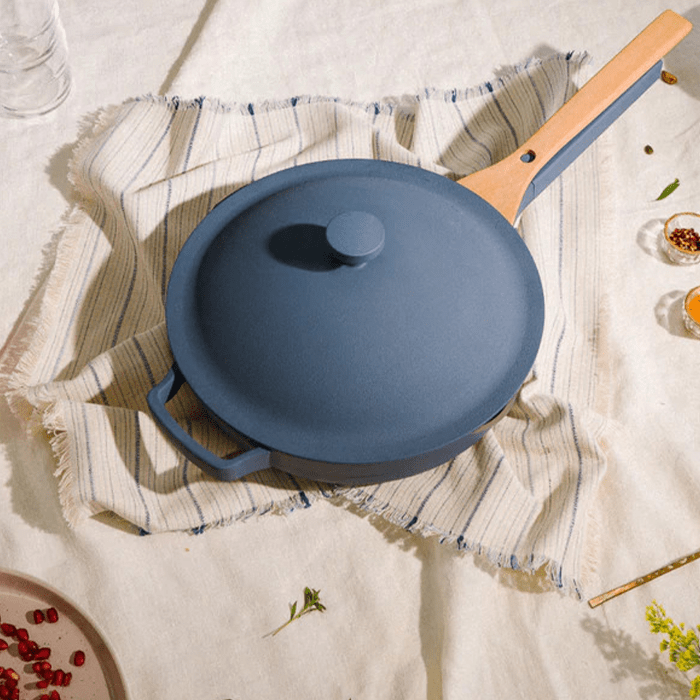 A blue frying pan with a wooden handle rests on a striped cloth, surrounded by scattered herbs and spices on a beige surface.