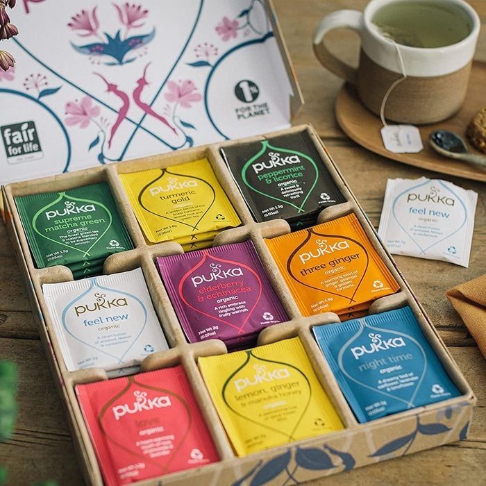 Tea bags arranged in a cardboard box; various flavors. Context includes a cup of tea, box lid with floral design, and wooden table.