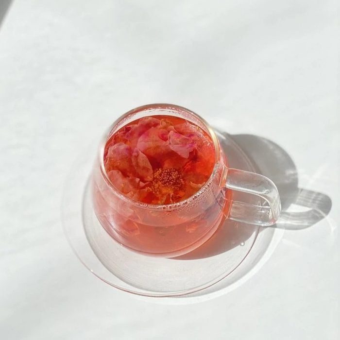 A clear glass cup holds pink flower tea under sunlight, casting a soft shadow on a white surface.