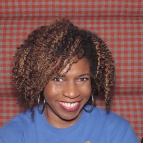 A person smiles brightly, sitting against a red plaid background, wearing a blue shirt and hoop earrings.