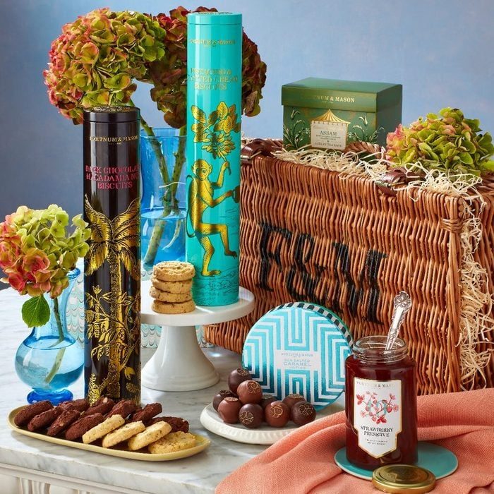 Assorted gourmet biscuits and jam displayed on a marble table with flowers, wicker hamper, and chocolates in a decorative setting.