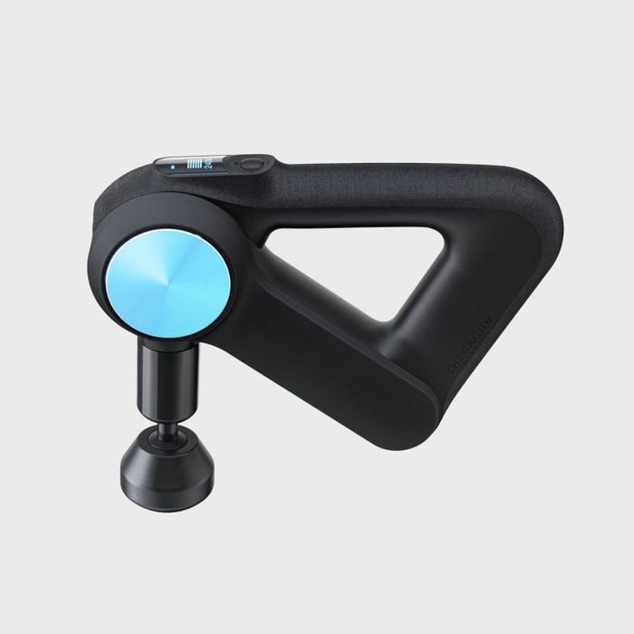 A black handheld massage gun with a triangular grip protrudes forward, featuring a metallic blue circular element. It is isolated on a gray background.