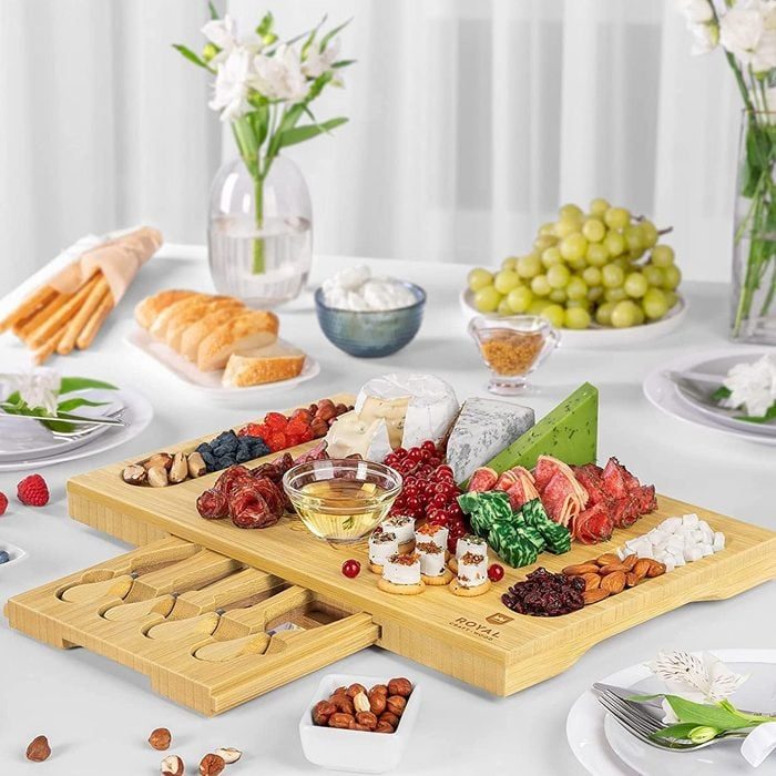 Cheese board displays assorted cheeses, meats, and fruits. Surrounding are fresh bread, grapes, and flowers on a white table with cutlery drawers.