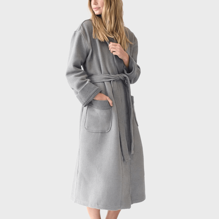 Woman wearing gray waffle-textured bathrobe, standing with hand in pocket, against a plain white background.