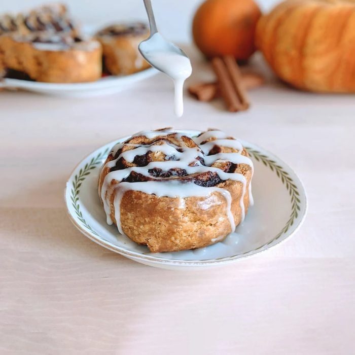 Icing drips onto a cinnamon roll on a plate, surrounded by pumpkins and cinnamon sticks on a wooden table.