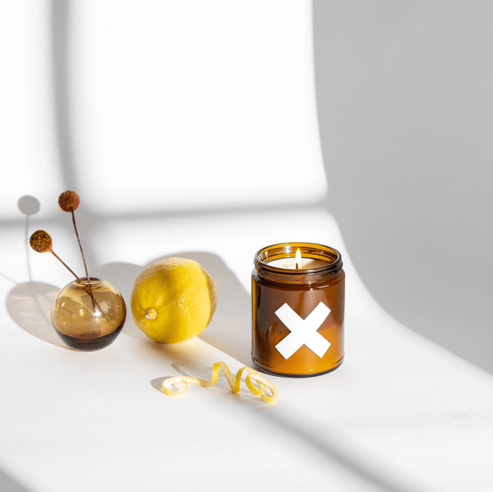 Brown candle burns beside a vase and lemon on a shadowed, white surface.