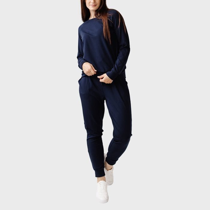 A person stands wearing a navy blue tracksuit and white sneakers, hands in pockets, against a plain gray background.