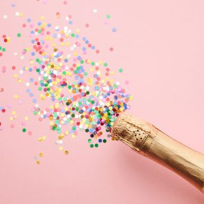 Champagne bottle releases colorful confetti onto a pink surface.