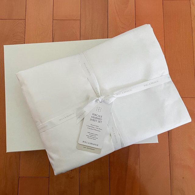 White sheet set lies folded, tied with branded ribbon on wood floor, labeled 