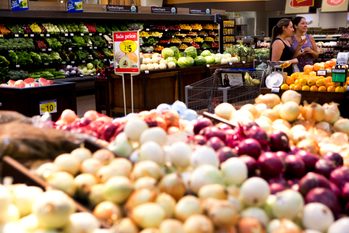 Onions displayed on tables, foreground; two people shopping, background; context is a supermarket produce section. Sign reads: "Sale price, Extra Large Limes, 2 for $5".