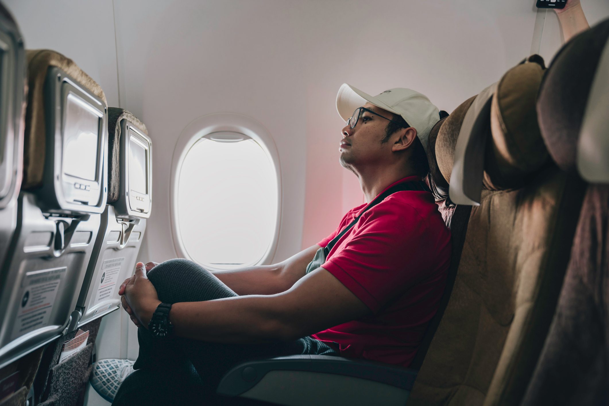 Man in a red shirt and white hat sitting in a window seat on an airplane