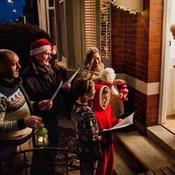 Group sings carols holding lanterns; an elderly woman smiles at open doorway on a residential street, illuminated by festive lights.