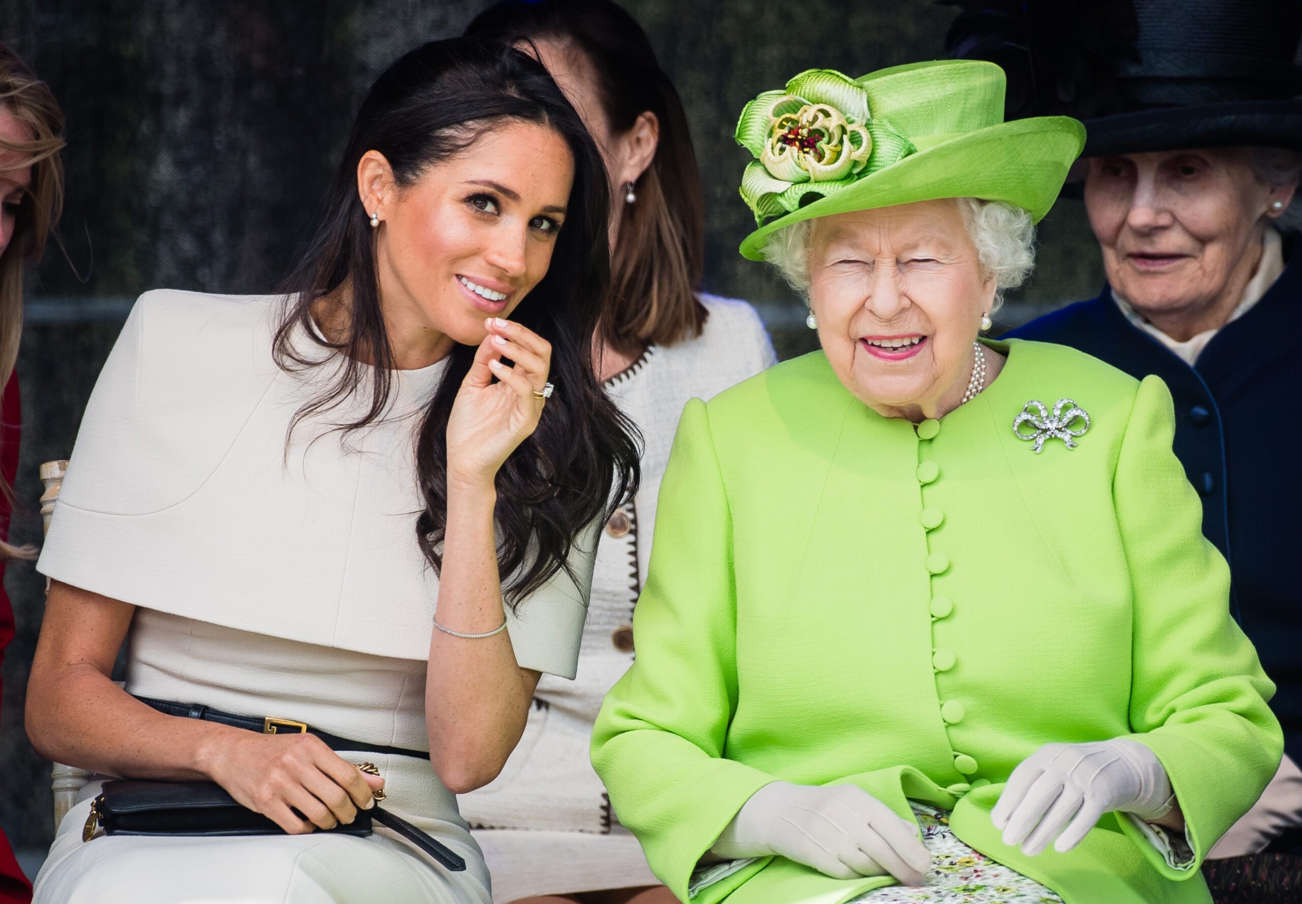 Two women sit together, smiling, in a public event. One wears white, and the other is in a bright green coat with a hat.