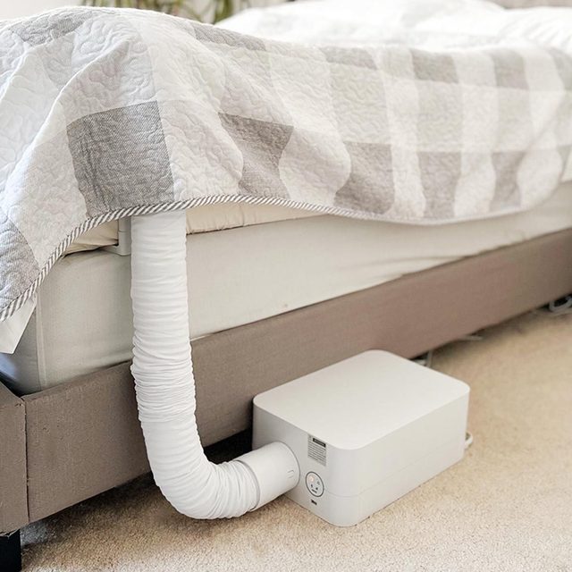 White hose extends into bed, connected to a white device on a carpeted floor, under a gray and white checkered blanket.