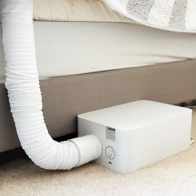 A white device connects a hose to a bed, resting on a carpeted floor in a bedroom.