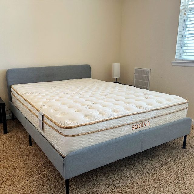 A mattress rests on a modern bed frame in a carpeted room with a small lamp near a window. Text on mattress: 