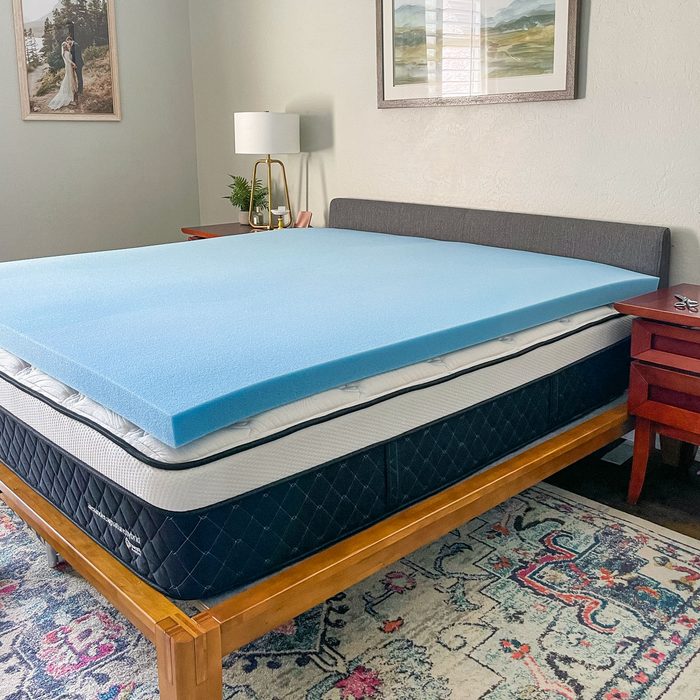 Mattress topper rests on a bed in a decorated room with a lamp, paintings, and a patterned rug.