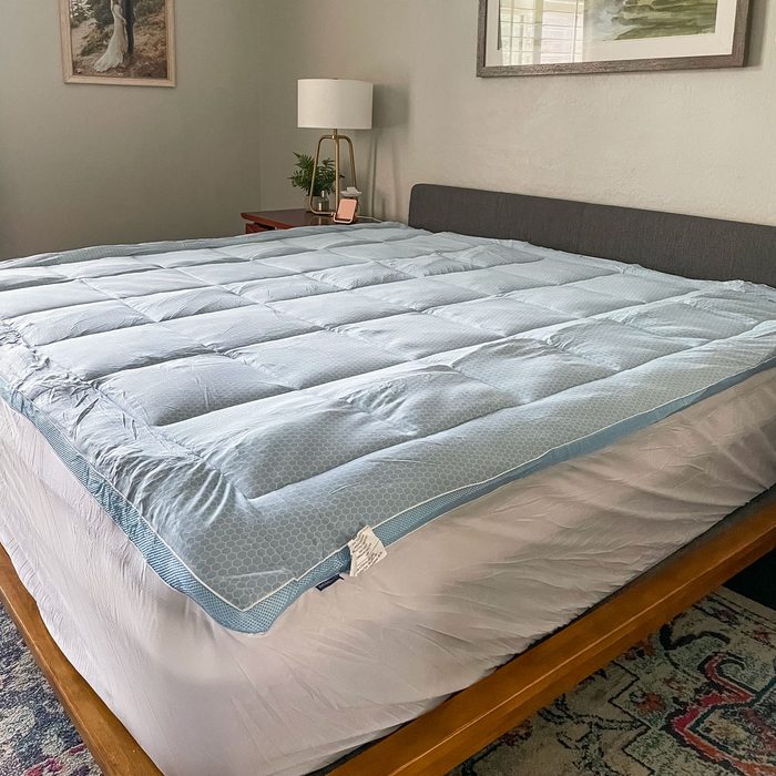 A thick, quilted mattress topper covers a bed in a cozy bedroom with framed art, a bedside lamp, and a patterned rug.