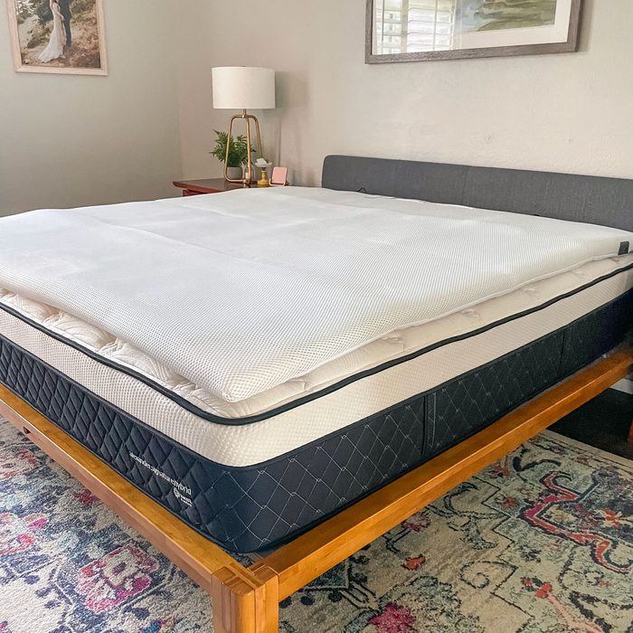 A mattress with a white topper rests on a wooden bed frame in a cozy bedroom with a lamp and artwork on the walls.