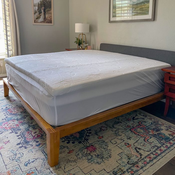 A neatly made bed with a white mattress sits on a wooden frame in a bedroom, featuring a rug, nightstand, and framed pictures.