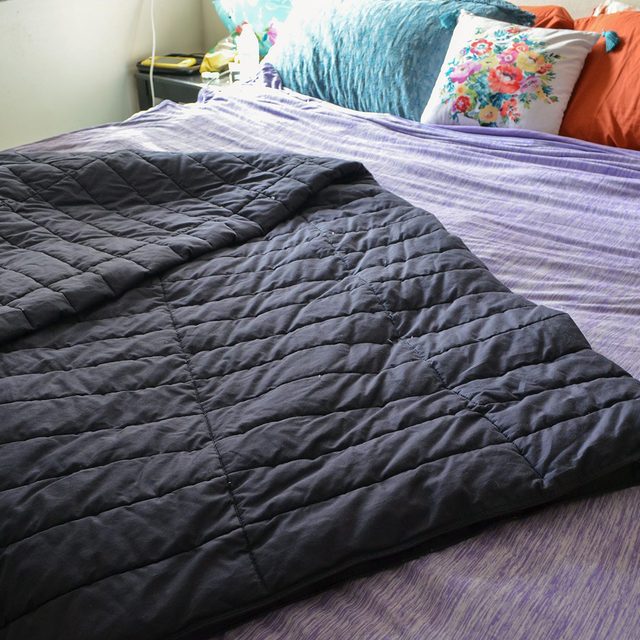 Quilted black blanket lies on a striped bed, surrounded by decorative pillows and a bedside table in a cozy bedroom.