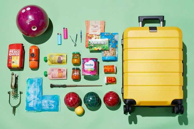 An array of odd items that are allowed in carryon bags arranged on a green background next to a carryon suitcase