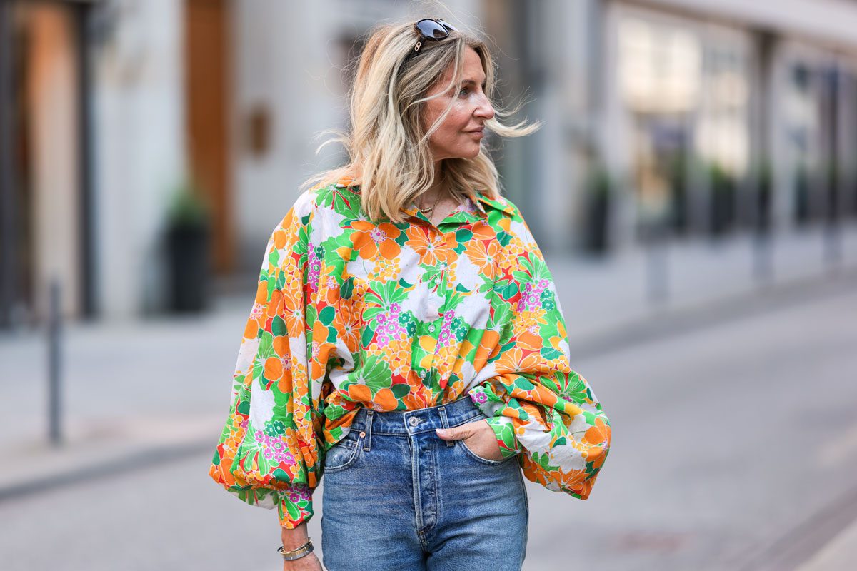 A person wearing a colorful, floral blouse and jeans walks along a city street, with sunglasses resting on their head.