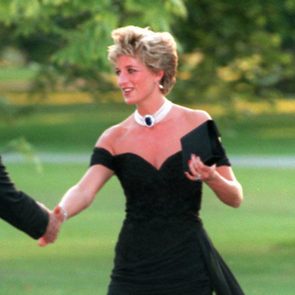 A person in a black dress shakes hands, outdoors, holding a black item, surrounded by greenery.